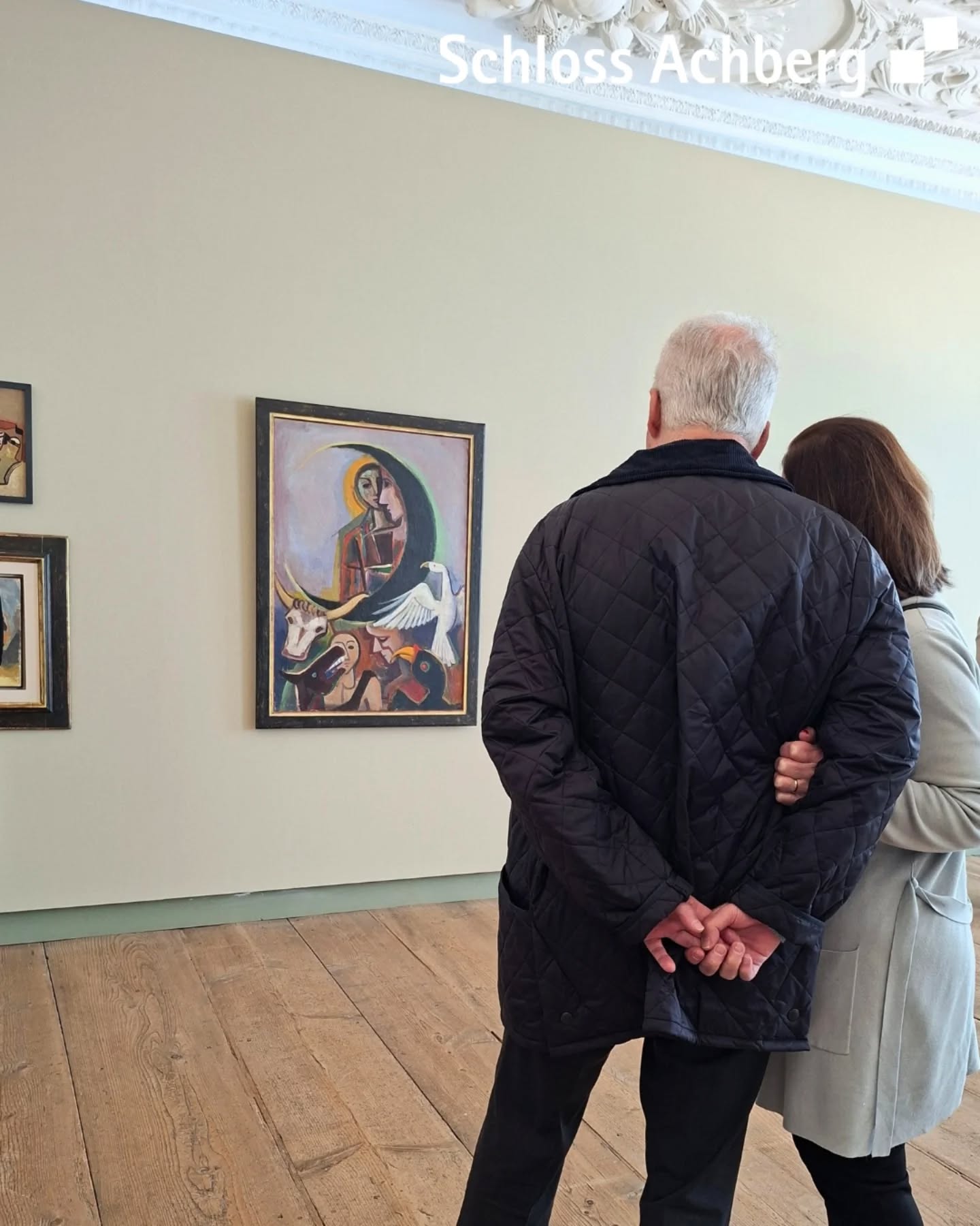 Dieser Alternativtext wurde KI-generiert: Zwei Personen betrachten ein Gemälde in einem hellen Raum. Das Bild zeigt eine abstrakte Darstellung mit Figuren und Farben.