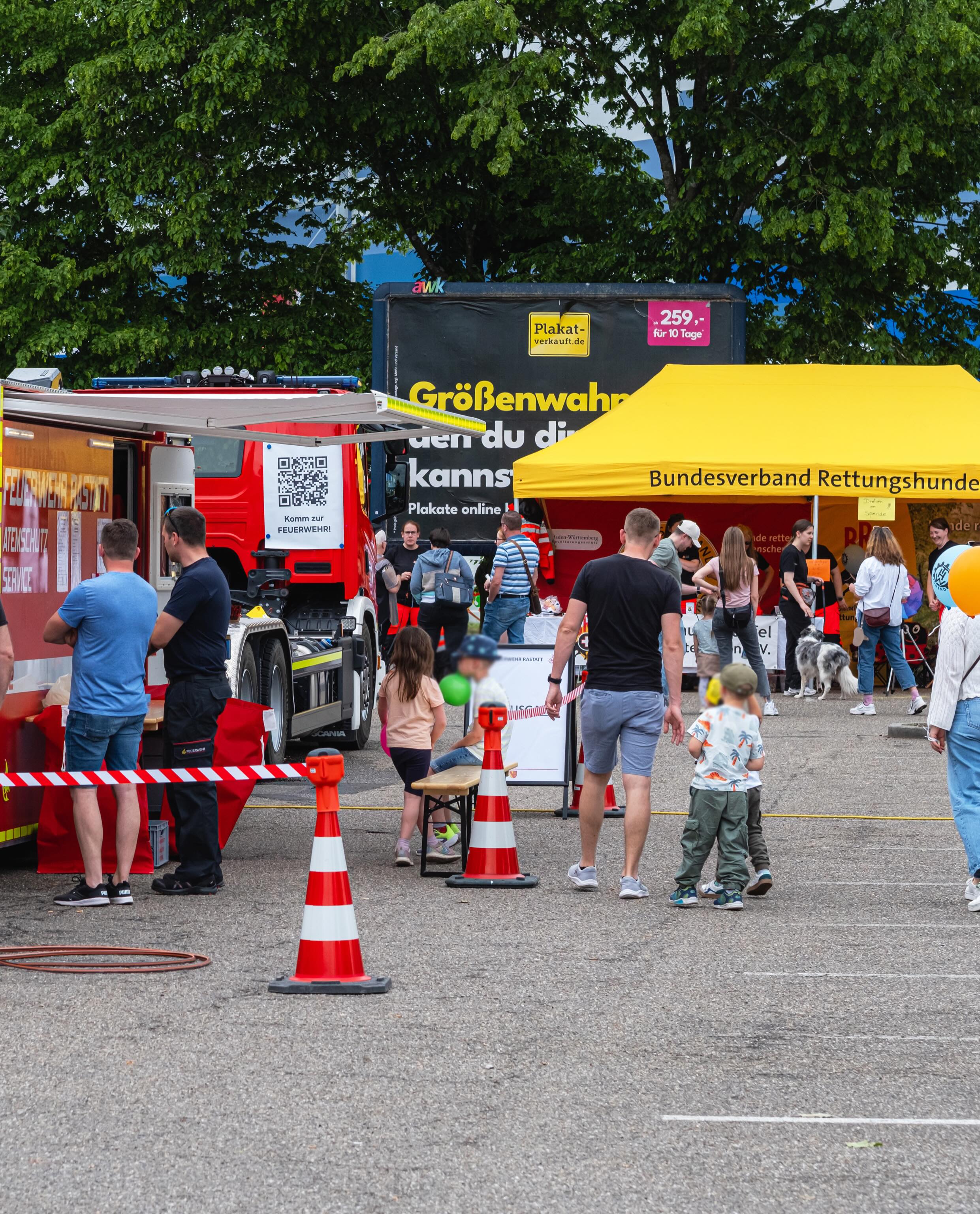 Dieser Alternativtext wurde KI-generiert: Eine rote Feuerwehrauto steht auf einer Straße, umgeben von Menschen und orangefarbenen Absperrhütchen. Im Hintergrund sind Plakate und Bäume zu sehen.