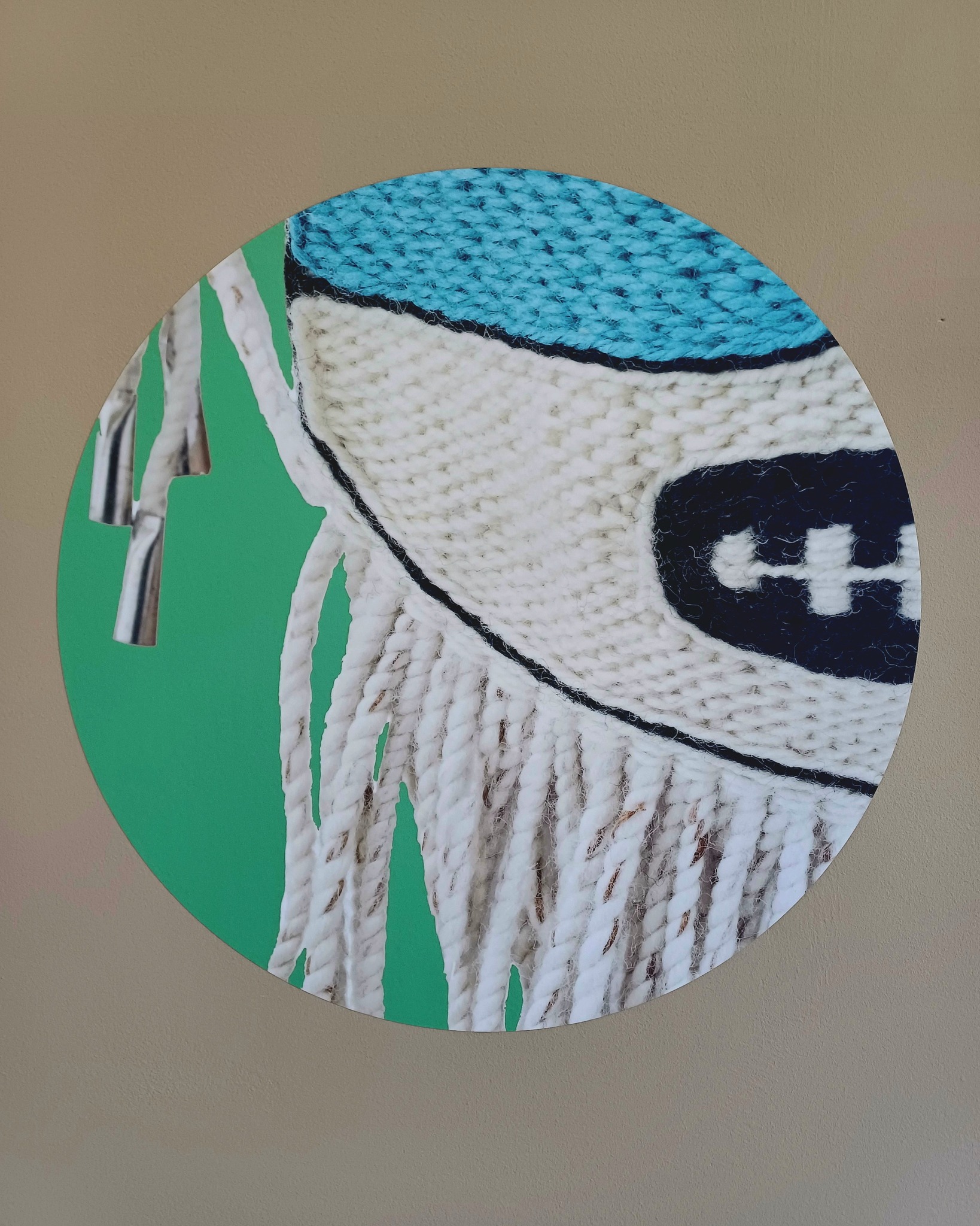Ein rundes Bild auf einer Wand zeigt eine Nahaufnahme eines gestrickten Objekts mit blauer Oberseite und weißem Mittelteil, auf dem ein dunkelblaues Logo zu sehen ist. Unten hängen weiße Fransen herab, während links silberne Streifen vor einem grünen Hintergrund platziert sind. (KI-Alternativtext)