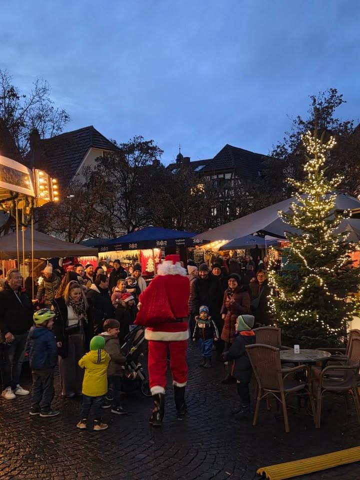 Dieser Alternativtext wurde KI-generiert: Ein belebter Platz mit Marktständen und einem festlich geschmückten Baum in der Dämmerung. Im Vordergrund ist eine Person in einem roten Kostüm zu sehen, umgeben von Menschen.
