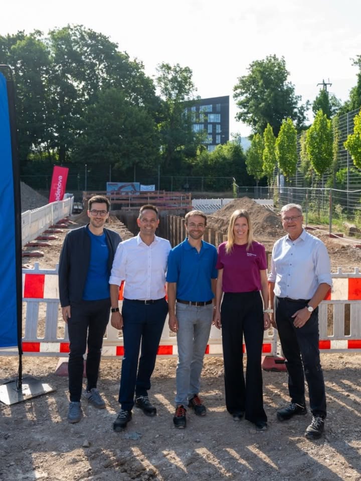 Fünf Personen stehen lächelnd nebeneinander auf einer Baustelle vor einer rot-weißen Absperrung. Im Hintergrund sind Bäume und ein Gebäude zu sehen, links steht ein rotes Banner mit der Aufschrift "solarova". (KI-Alternativtext)