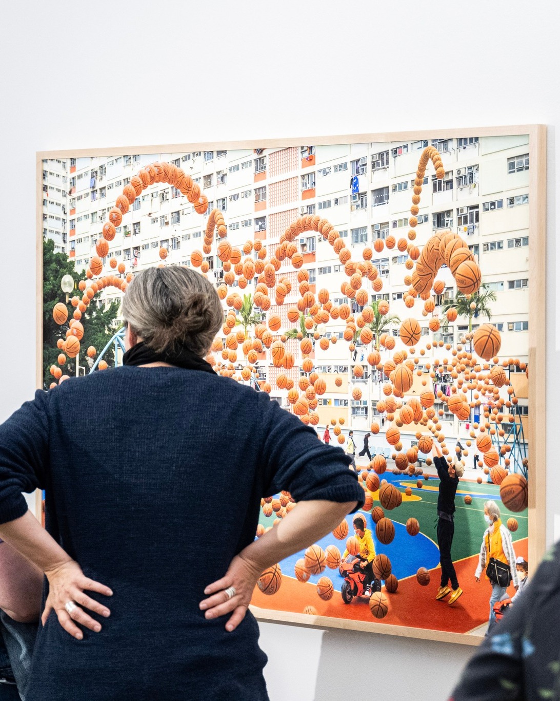 Eine Person in einem dunklen Pullover steht mit Händen in den Hüften vor einem großen, gerahmten Bild. Das Kunstwerk zeigt einen Basketballplatz vor einem Hochhaus, über dem schwebende Bälle das Wort „HAPPY“ formen. Auf dem Platz sind weitere Personen zu sehen, darunter eine, die nach oben greift, und eine auf einem kleinen roten Fahrzeug. (KI-Alternativtext)