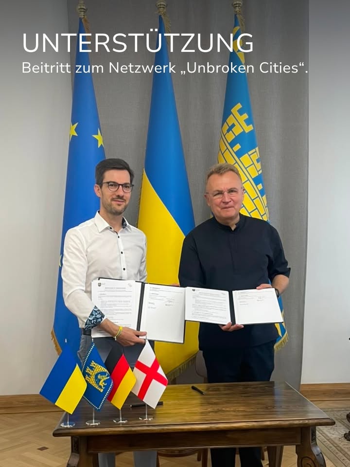 Zwei Personen halten vor drei großen Flaggen (EU, Ukraine, eine weitere Flagge) offene Dokumente. Auf dem Tisch davor stehen kleine Flaggen der Ukraine, der weiteren Flagge, Deutschlands und Englands. Der Text im Bild lautet: „UNTERSTÜTZUNG Beitritt zum Netzwerk ‚Unbroken Cities‘.“ (KI-Alternativtext)