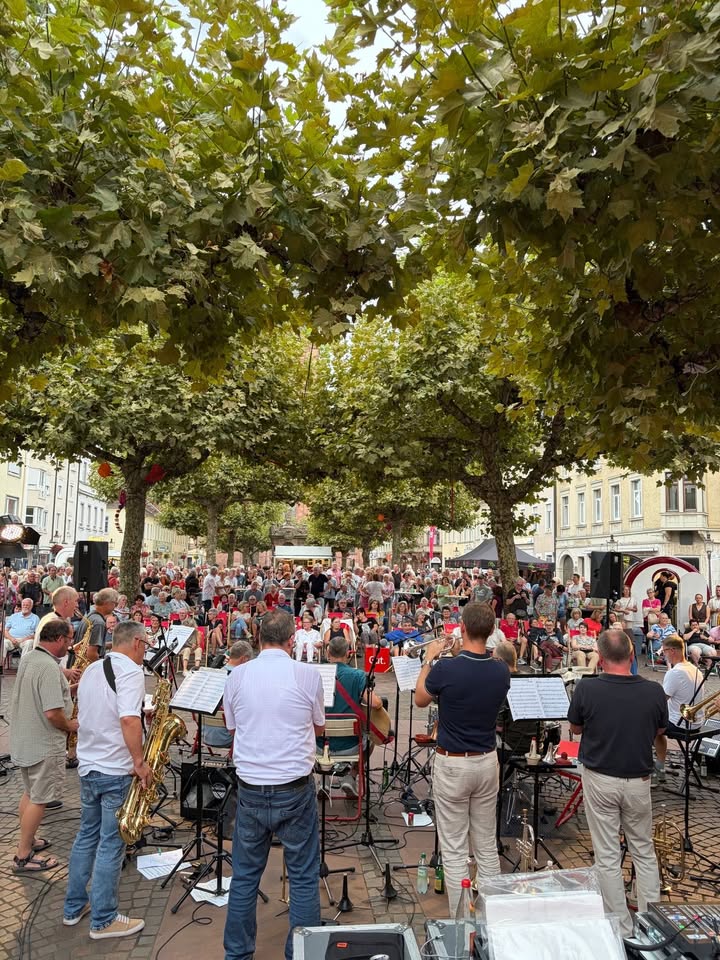 Mehrere Musiker stehen mit Blasinstrumenten vor Notenständern und spielen im Freien. Vor ihnen sitzt und steht ein großes Publikum auf einem von Bäumen gesäumten Platz. Im Hintergrund sind städtische Gebäude zu sehen. (KI-Alternativtext)