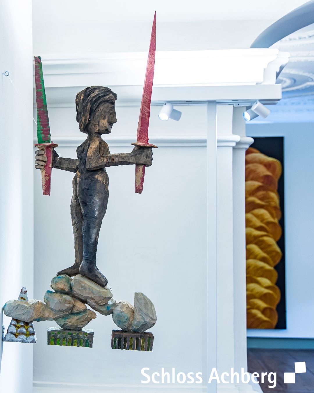 Eine skulpturale Figur hält zwei lange, schmale Objekte. Die Szene befindet sich in einem hellen Raum, möglicherweise einer Ausstellung.