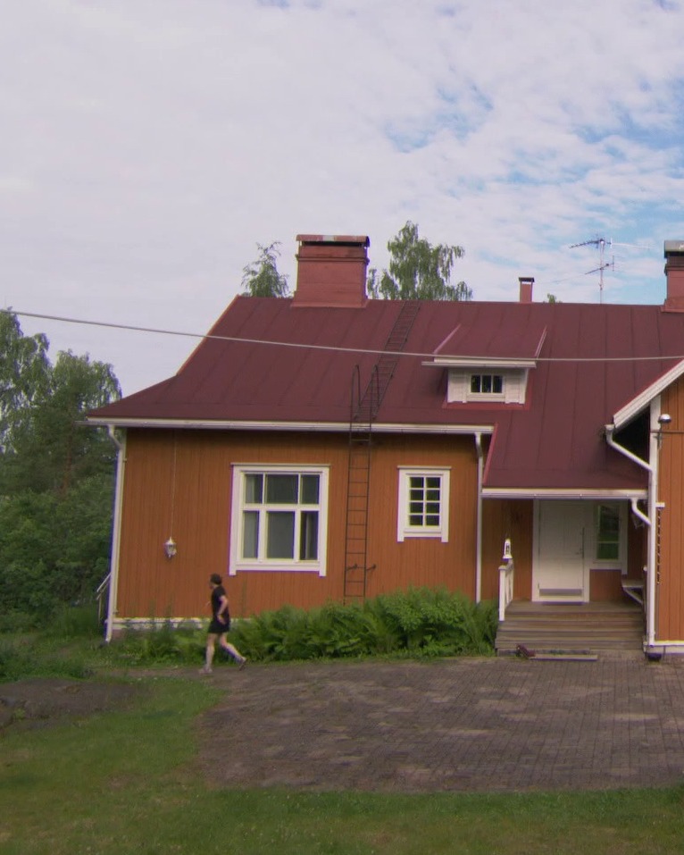 Ein orangefarbenes Holzhaus mit rotem Dach und weißen Fensterrahmen. Eine Leiter lehnt an der Dachseite. Vor dem Haus läuft eine Person von links nach rechts vorbei, während grüne Büsche die Fassade säumen. (KI-Alternativtext)
