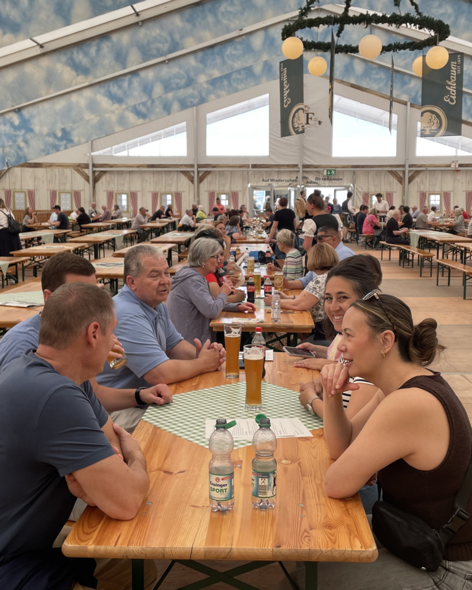 Menschen sitzen an langen Holztischen in einem großen Zelt mit blau-weißem Himmelsgemälde an der Decke und trinken Bier und Wasser. An den Wänden hängen Werbebanner, darunter eines mit „Eichbaum“. Im Hintergrund sind weitere Gäste und ein Ausgang mit der Aufschrift „Auf Wiedersehen“ zu sehen. (KI-Alternativtext)