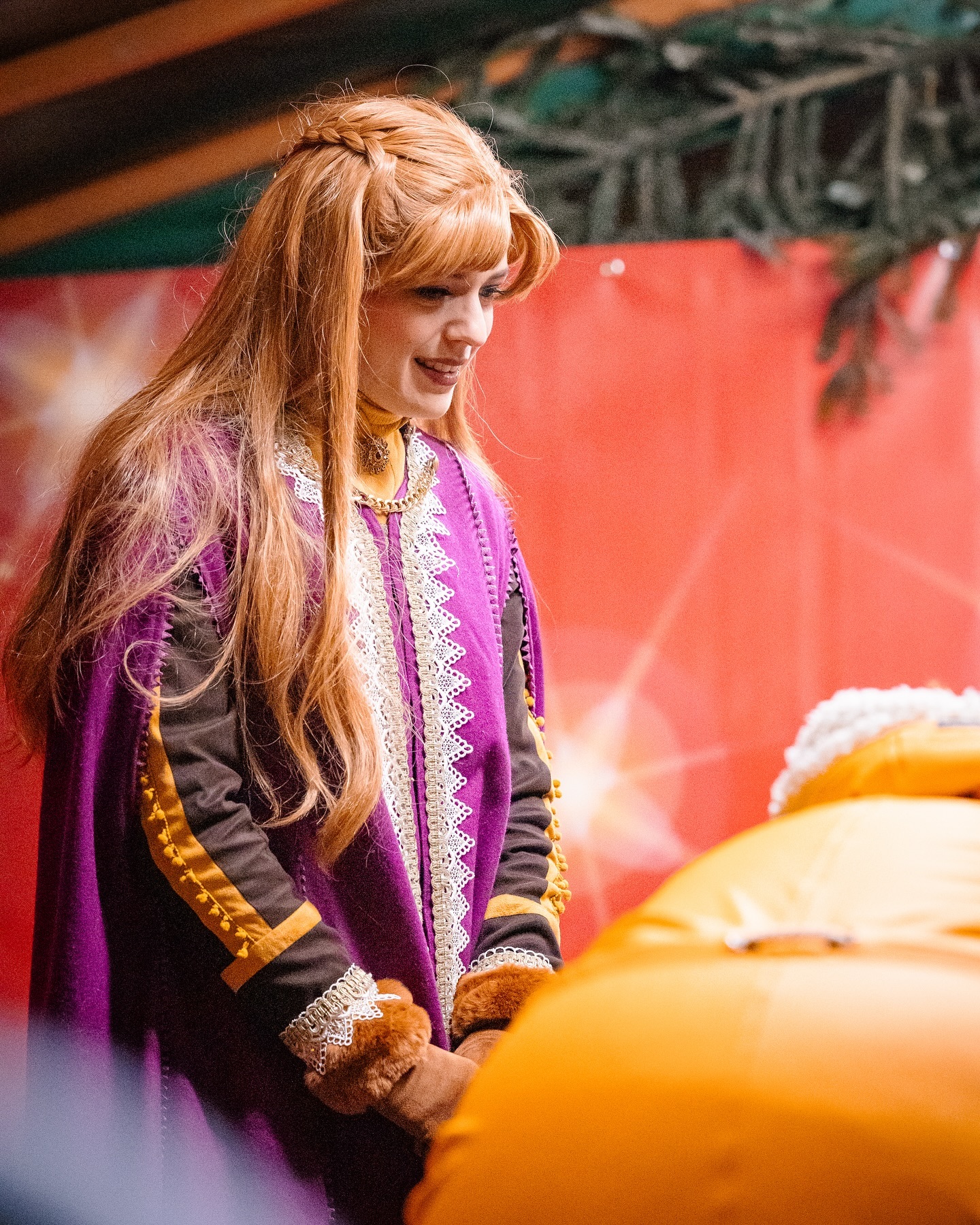 Eine Person in einem violetten Gewand mit goldenen Details steht vor einem orangenen Gegenstand. Das Haar ist rot und zu einem Zopf geflochten.