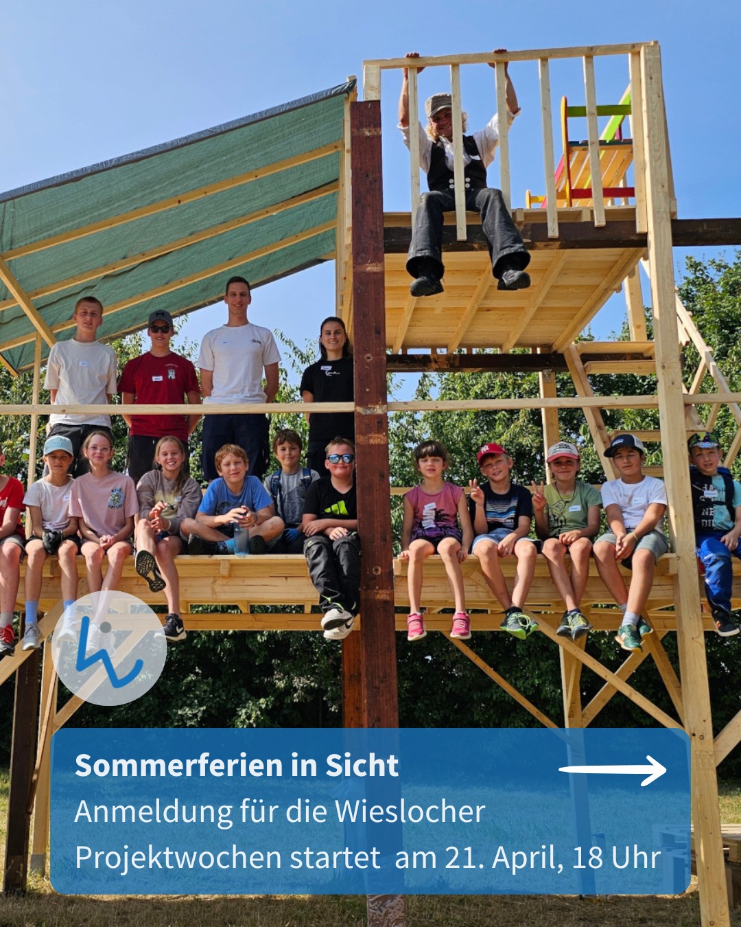 Dieser Alternativtext wurde KI-generiert: Eine Gruppe von Personen steht und sitzt vor einer Holzkonstruktion im Freien. Im Hintergrund ist ein Himmel zu sehen, das Bild ist farbig.