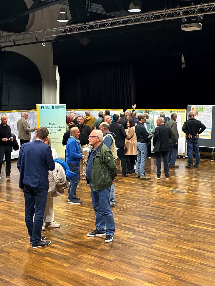 Dieser Alternativtext wurde KI-generiert: Eine Gruppe von Personen steht in einer großen Halle mit Tischen und Plakaten. Im Hintergrund befinden sich dunkle Vorhänge und ein Bühnenbereich.