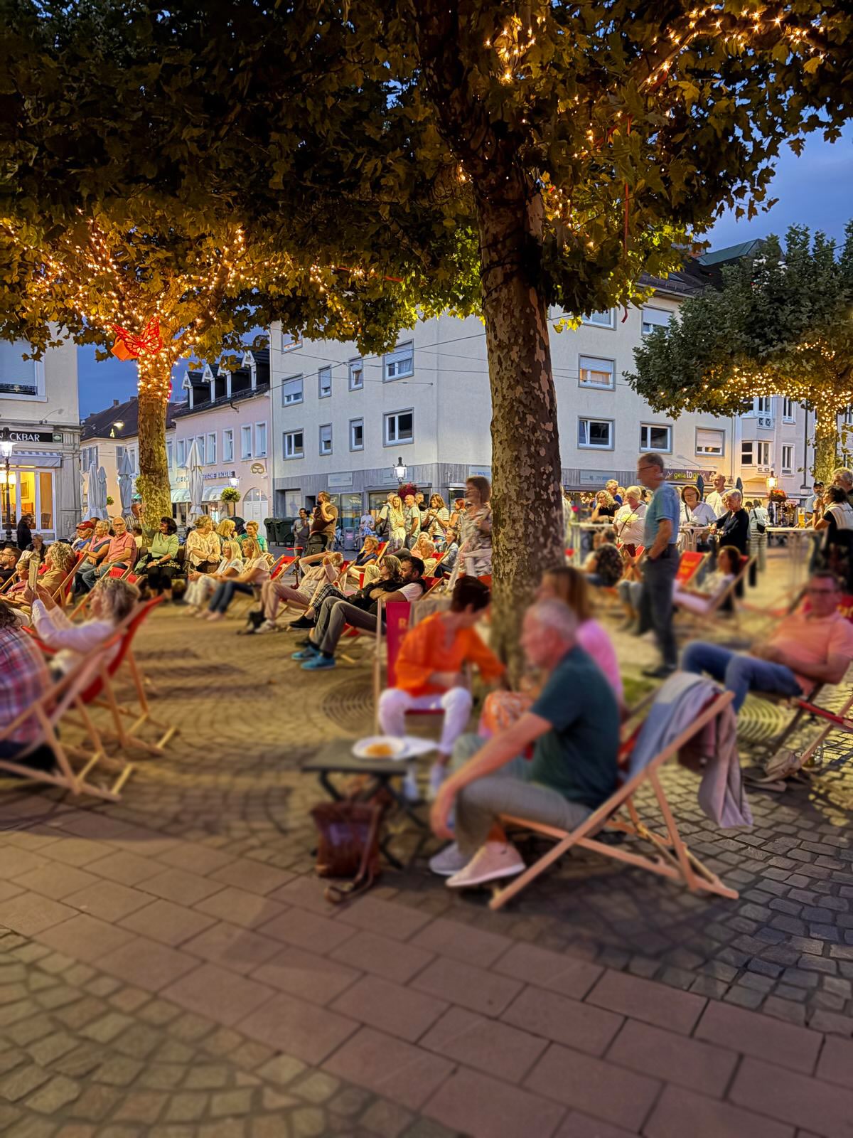 Eine große Gruppe von Menschen sitzt abends auf einem gepflasterten Platz in Reihen von Liegestühlen. Bäume mit Lichterketten und beleuchtete Gebäude im Hintergrund deuten auf ein Open-Air-Event hin. (KI-Alternativtext)