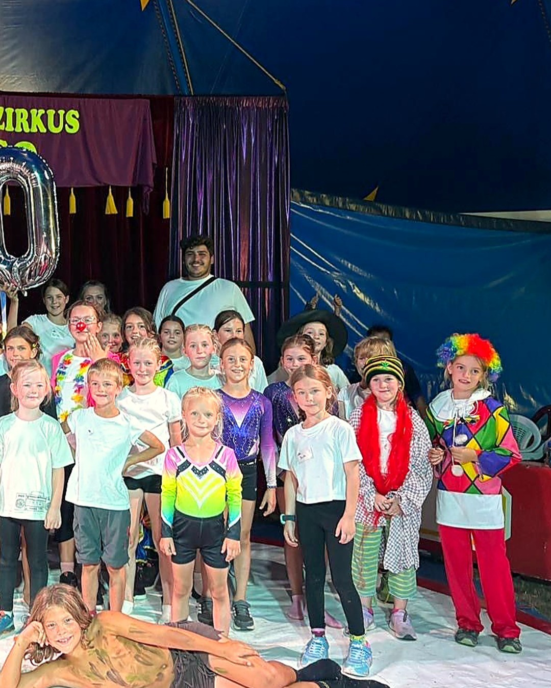 Eine große Gruppe von Kindern und eine erwachsene Person posieren vor einem blauen Zelt mit einem Schild, das „ZIRKUS“ besagt. Viele der Kinder tragen bunte Kostüme, wie Clown-Kleidung, Turnanzüge oder Kopfbedeckungen, während ein Kind im Vordergrund mit Gesichtsbemalung auf dem Boden liegt. Links schwebt ein silberner Folienballon in der Form einer Null. (KI-Alternativtext)