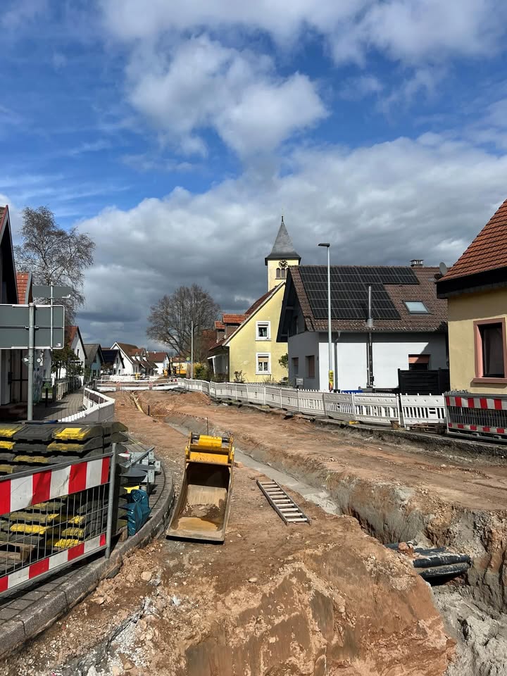 Dieser Alternativtext wurde KI-generiert: Eine Aufnahme einer Straßenszene mit Bauarbeiten im Vordergrund. Im Hintergrund sind Gebäude mit roten Dächern und ein Kirchturm zu sehen.