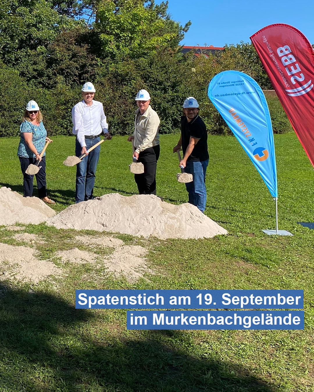 Dieser Alternativtext wurde KI-generiert: Eine Gruppe von Personen mit Schutzhelmen und Westen hält Schaufeln in der Hand und steht auf einer Baustelle. Im Vordergrund ein Banner mit Aufschrift und Datum.