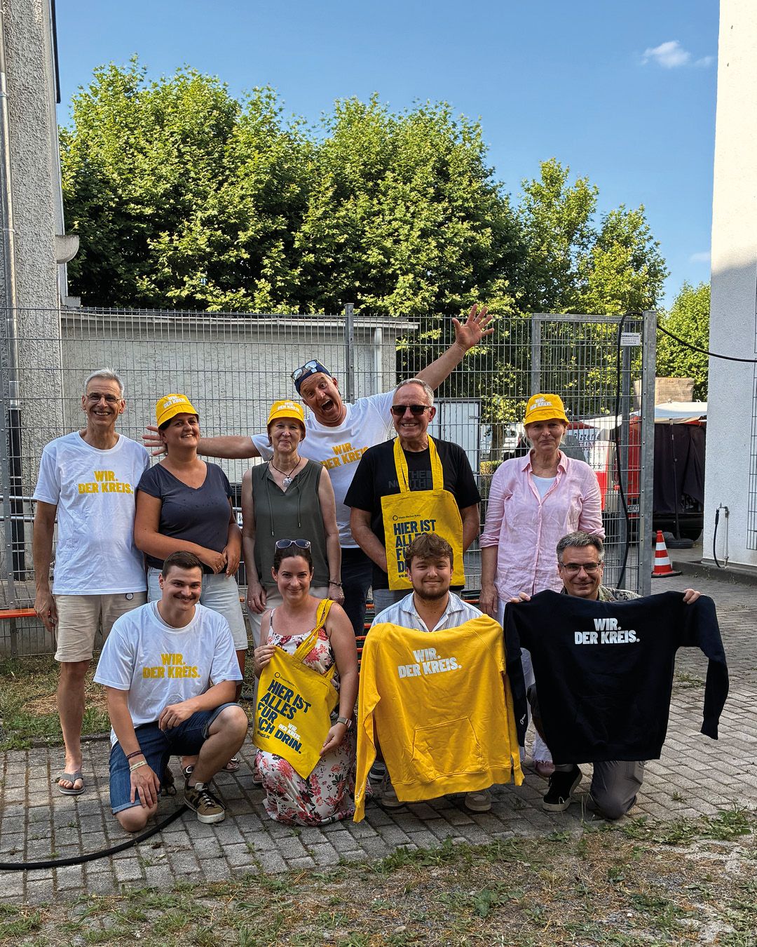 Eine Gruppe von zehn Personen posiert im Freien vor einem Metallzaun und Bäumen. Mehrere tragen weiße T-Shirts oder gelbe Kopfbedeckungen mit der Aufschrift „WIR. DER KREIS.“. Einige halten gelbe und schwarze Kleidungsstücke sowie eine Tasche mit dem Text „HIER IST ALLES FÜR DICH DRIN.“ in die Höhe. (KI-Alternativtext)