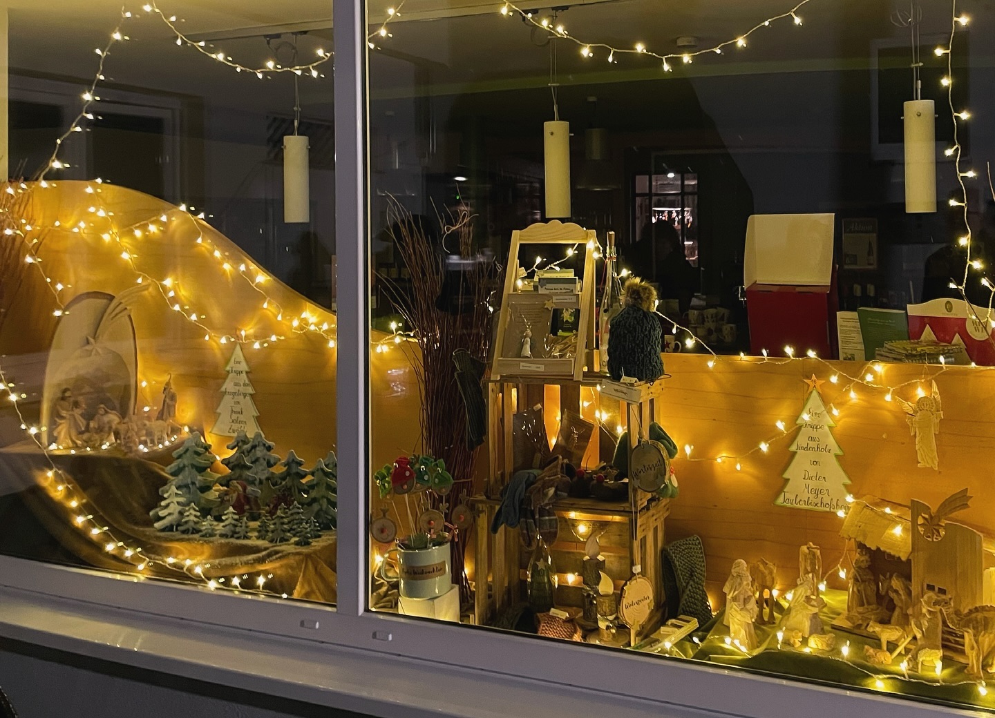 Festlich geschmücktes Schaufenster mit Weihnachtskrippe, Lichtern und weihnachtlichen Dekorationen. Es sind verschiedene Artikel zu sehen.