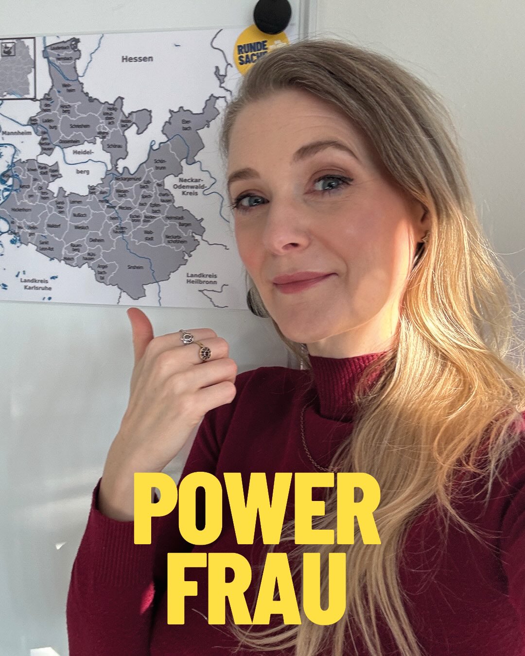 Dieser Alternativtext wurde KI-generiert: Eine Person steht vor einer Karte und hält die Hände hoch. Im Hintergrund ist ein Fenster zu sehen und gelber Text mit der Aufschrift „POWER FRAU“.