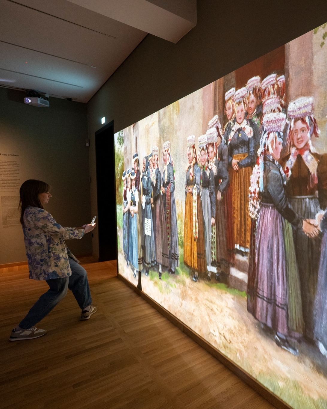 Dieser Alternativtext wurde KI-generiert: Eine Person betrachtet ein großes, projiziertes Gemälde in einem Museum. Das Gemälde zeigt eine Gruppe von Menschen in traditioneller Kleidung.