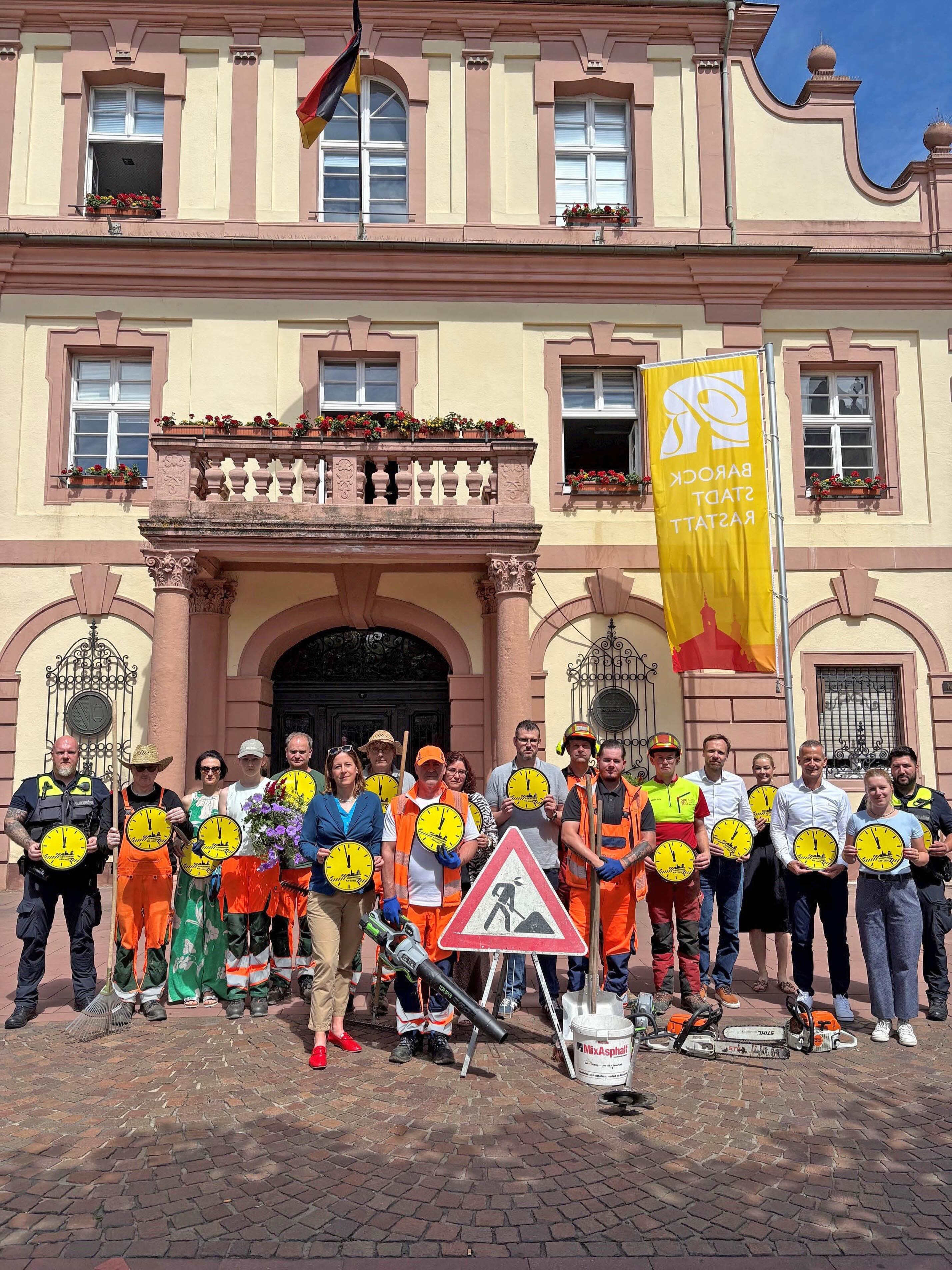 Gruppe von Menschen vor einem barocken Rathaus, die gelbe Uhren-Schilder halten und Baustellenutensilien wie Schilder, Werkzeuge und einen Eimer präsentieren; eine gelbe Fahne mit der Aufschrift „BAROCK STADT RASTATT“ ist rechts sichtbar. (KI-Alternativtext)