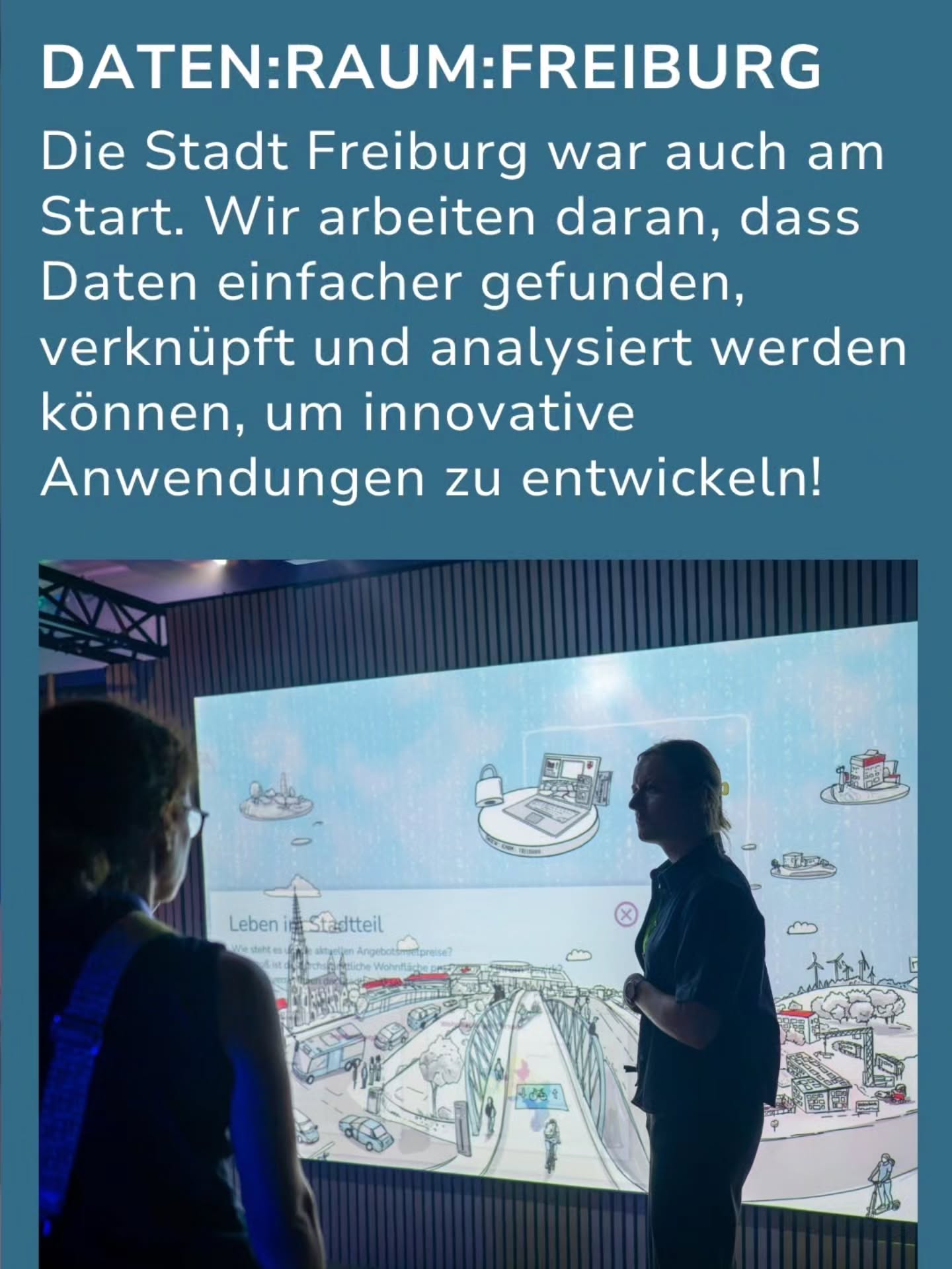 DATEN:RAUM:FREIBURG. Die Stadt Freiburg war auch am Start. Wir arbeiten daran, dass Daten einfacher gefunden, verknüpft und analysiert werden können, um innovative Anwendungen zu entwickeln! Zwei Personen stehen vor einer großen Projektionsfläche, auf der eine Illustration eines Stadtteils mit Daten-Symbolen zu sehen ist. (KI-Alternativtext)