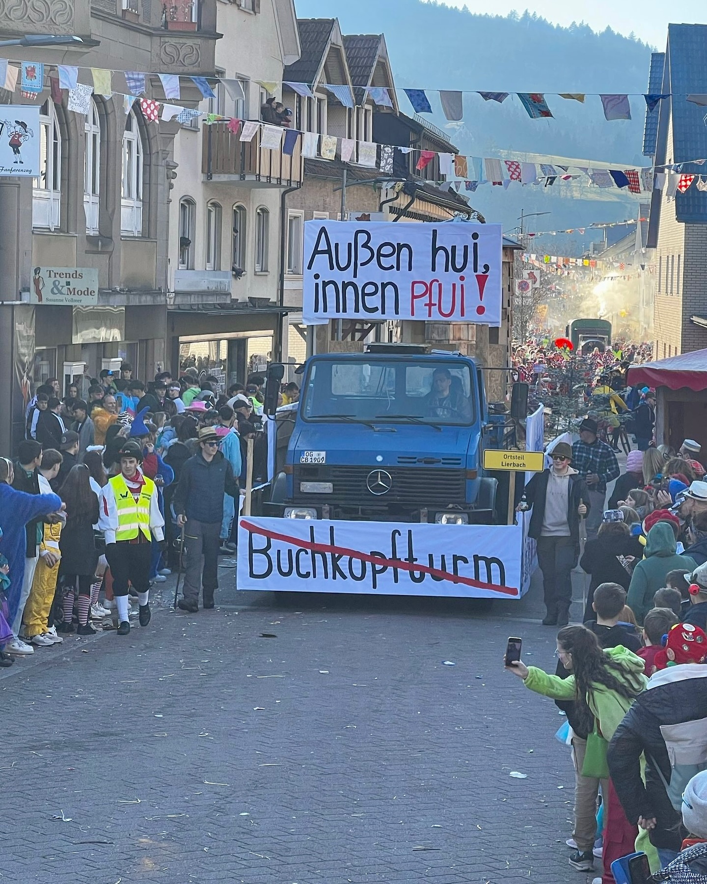 Ein Karnevalsumzug mit einem blauen LKW, auf dem Schilder befestigt sind. Viele Leute säumen die Straße, und im Hintergrund sind Häuser und eine Bergkette zu sehen.