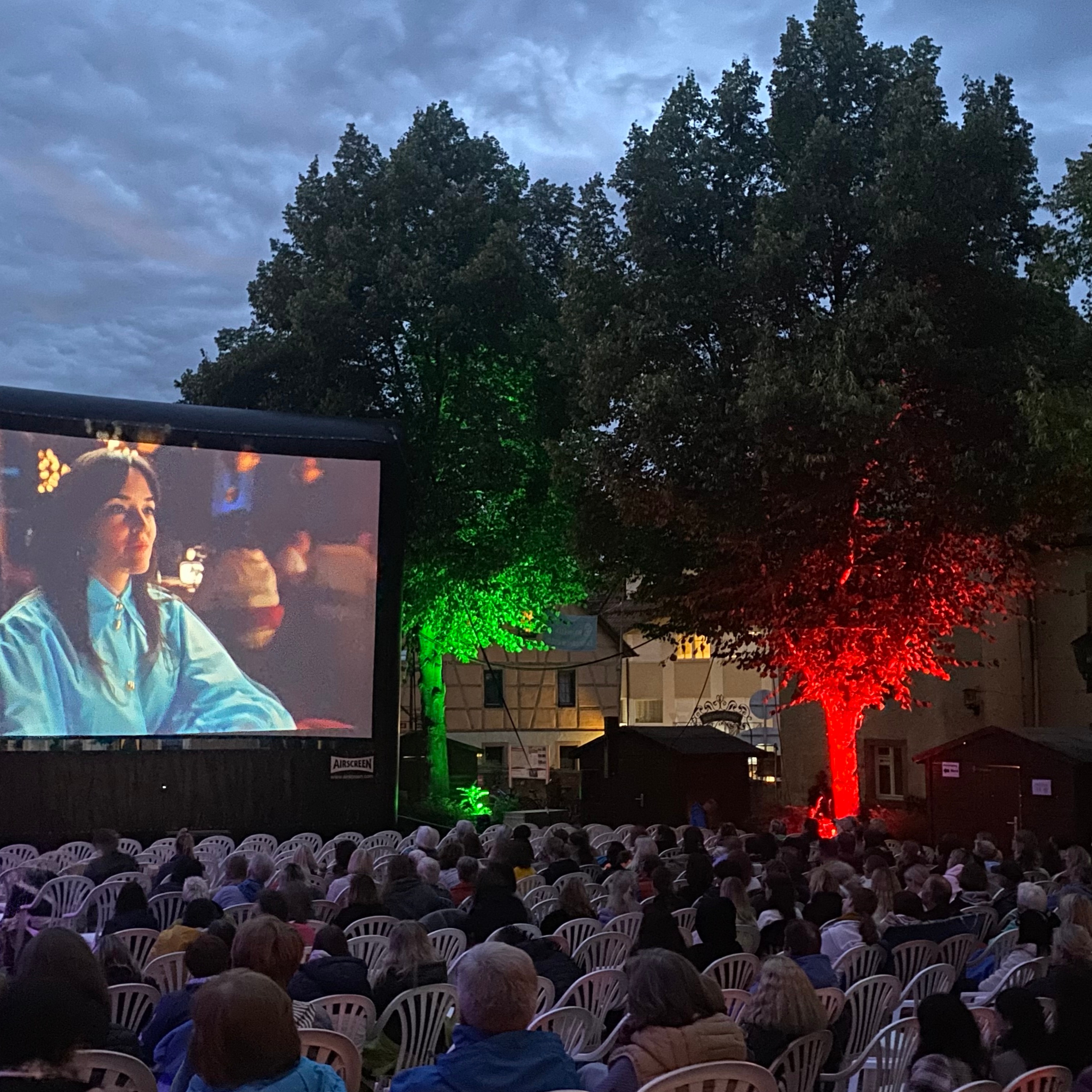Ein Open-Air-Kino bei Dämmerung. Auf der großen Leinwand links wird eine Person in einem blauen Hemd gezeigt. Eine große Menschenmenge sitzt auf weißen Stühlen und schaut auf den Bildschirm, während im Hintergrund Bäume in Grün und Rot beleuchtet sind. (KI-Alternativtext)