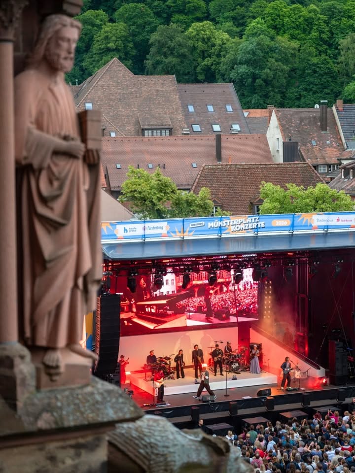 Statue im Vordergrund, dahinter Bühne mit Musikern bei einem Konzert vor Publikum; im Hintergrund Dächer und Bäume, Banner mit „Münsterplatz Konzerte 2024“. (KI-Alternativtext)