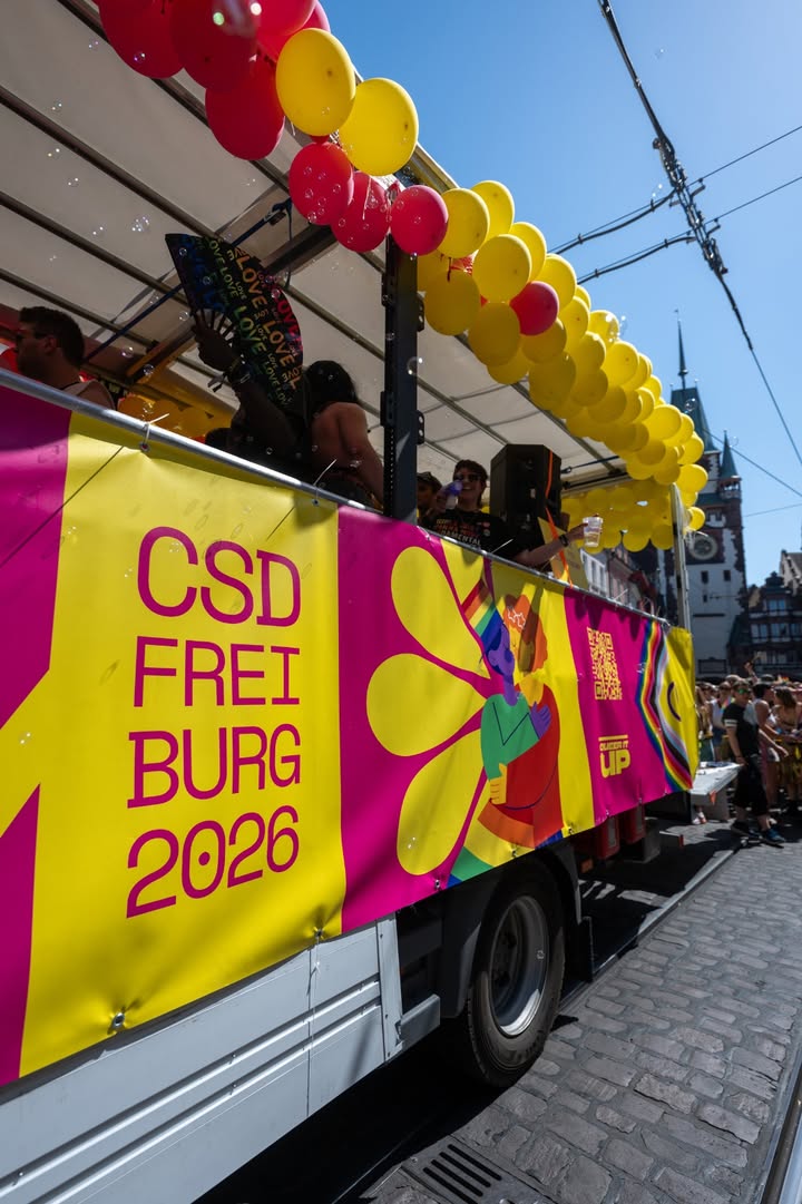 Ein Parade-Wagen mit gelb-pinkem Banner „CSD FREIBURG 2026“, geschmückt mit roten und gelben Luftballons. Personen stehen darauf, im Hintergrund eine historische Turmsilhouette unter blauem Himmel. (KI-Alternativtext)