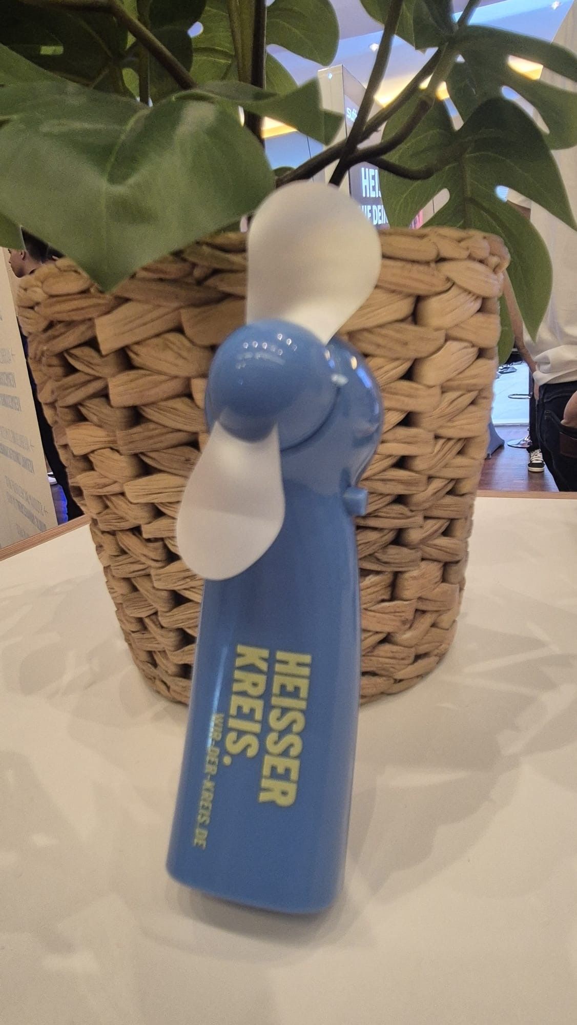 Ein blauer Handventilator mit weißen Flügeln steht im Vordergrund auf einer weißen Oberfläche. Auf dem Gerät ist in gelber Schrift „HEISSER KREIS.“ sowie die Webadresse „WIR-DER-KREIS.DE“ zu lesen. Dahinter befindet sich ein geflochtener Korb mit einer Pflanze mit großen grünen Blättern. (KI-Alternativtext)