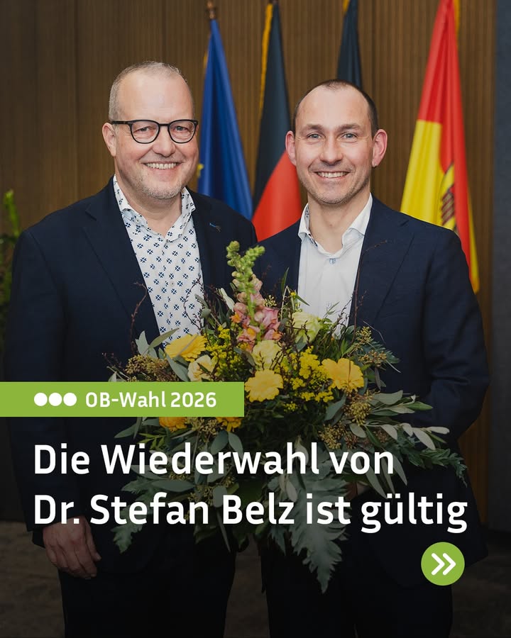 Dieser Alternativtext wurde KI-generiert: Zwei Personen stehen lächelnd nebeneinander und halten einen Blumenstrauß. Im Hintergrund sind Fahnen zu sehen.
