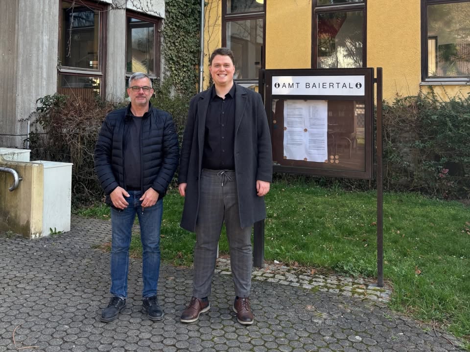 Dieser Alternativtext wurde KI-generiert: Zwei Personen stehen vor einem gelben Gebäude mit einem Informationstafel. Im Hintergrund sind Fenster und Büsche zu sehen.