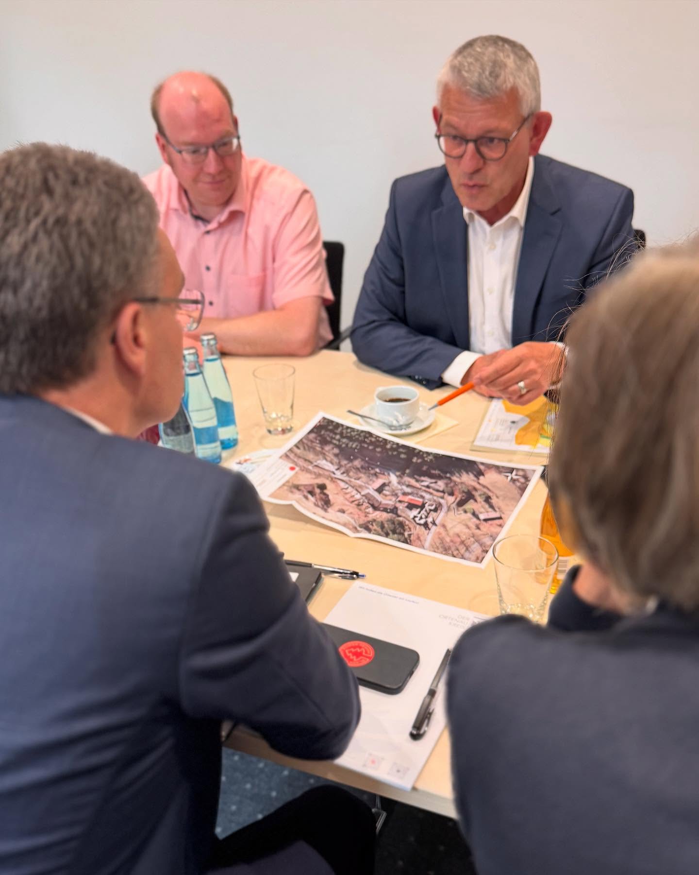 Eine Gruppe von Personen sitzt an einem Tisch und betrachtet ein Foto. Auf dem Tisch stehen Getränke, ein Handy und Unterlagen.