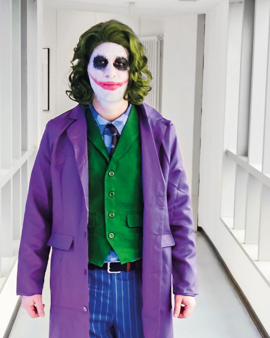 Dieser Alternativtext wurde KI-generiert: Eine Person trägt ein Kostüm mit grünem Haar, violettem Mantel und einem grünen Hemd. Die Person steht in einem hellen Gang.