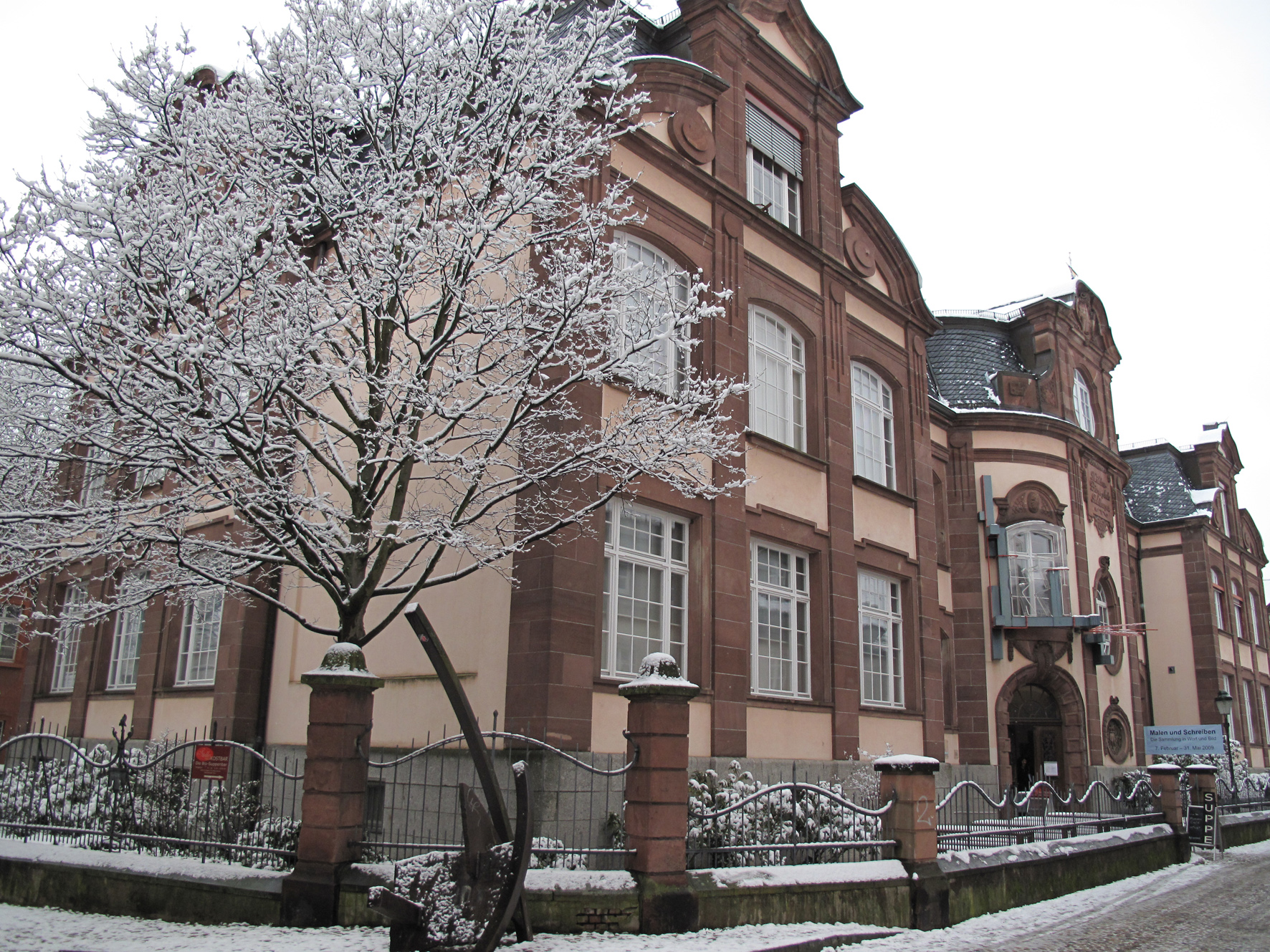 Dieser Alternativtext wurde KI-generiert: Ein Gebäude mit roten und beigen Fassadenelementen steht neben einem eisbedeckten Zaun und Baum. Schnee liegt auf dem Boden und den Ästen, was eine winterliche Atmosphäre erzeugt.