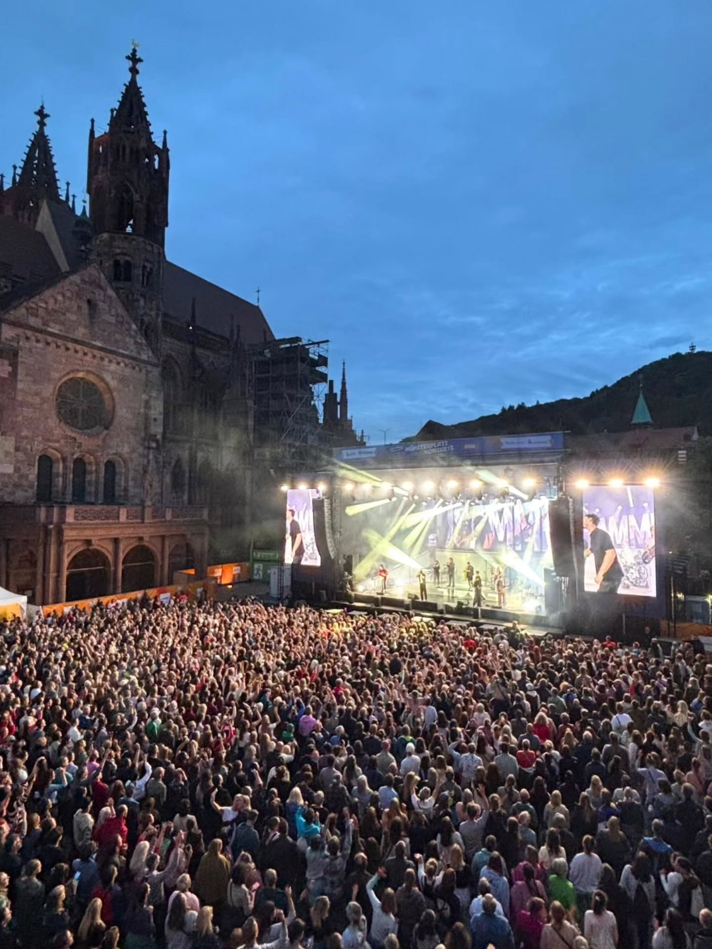 Große Menschenmenge vor einer beleuchteten Bühne bei einem Konzert im Freien, im Hintergrund eine gotische Kirche bei Dämmerung. Auf der Bühne sind Musiker und große Bildschirme mit Live-Übertragung zu sehen. (KI-Alternativtext)