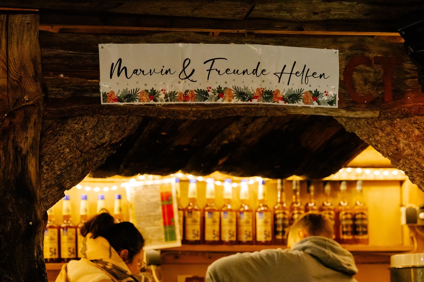Ein Schild mit weihnachtlicher Dekoration über einer Theke mit Flaschen. Im Hintergrund sind Menschen zu sehen.