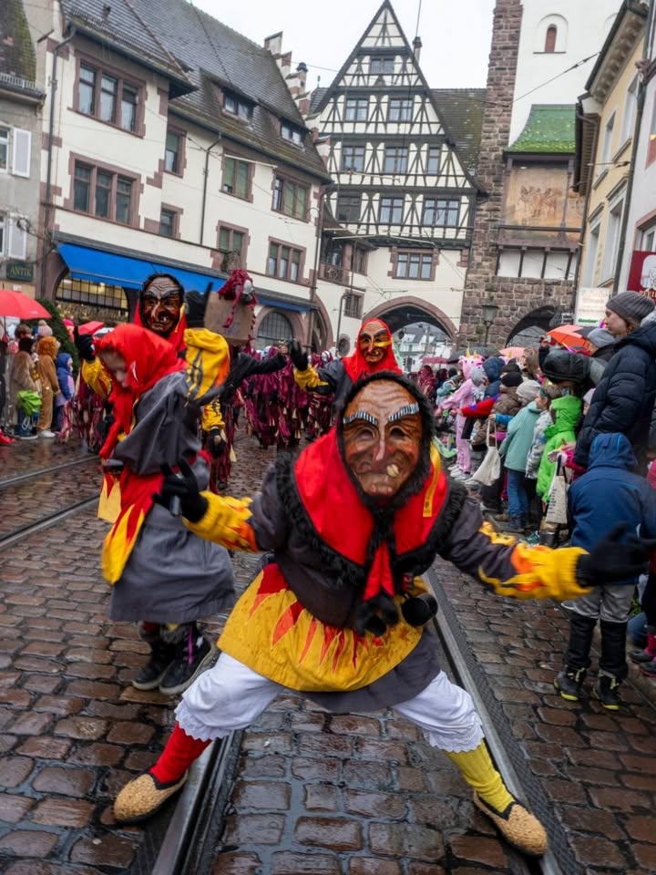 Dieser Alternativtext wurde KI-generiert: Ein Straßenbild mit traditionellen, farbenfrohen Masken und Figuren, die an einem Festzug teilnehmen. Im Hintergrund sind historische Fachwerkhäuser zu sehen.