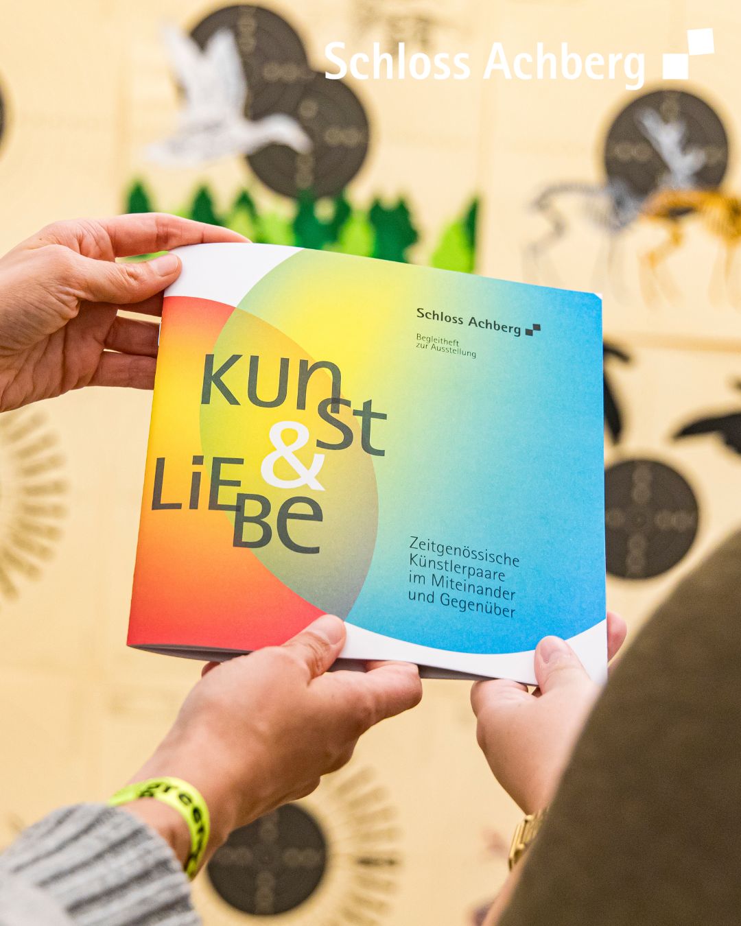 Eine Person hält ein farbiges Buch oder eine Broschüre in den Händen, auf dem Frontcover ist Text zu sehen. Im Hintergrund sind Pflanzen und ein Gebäude angedeutet.