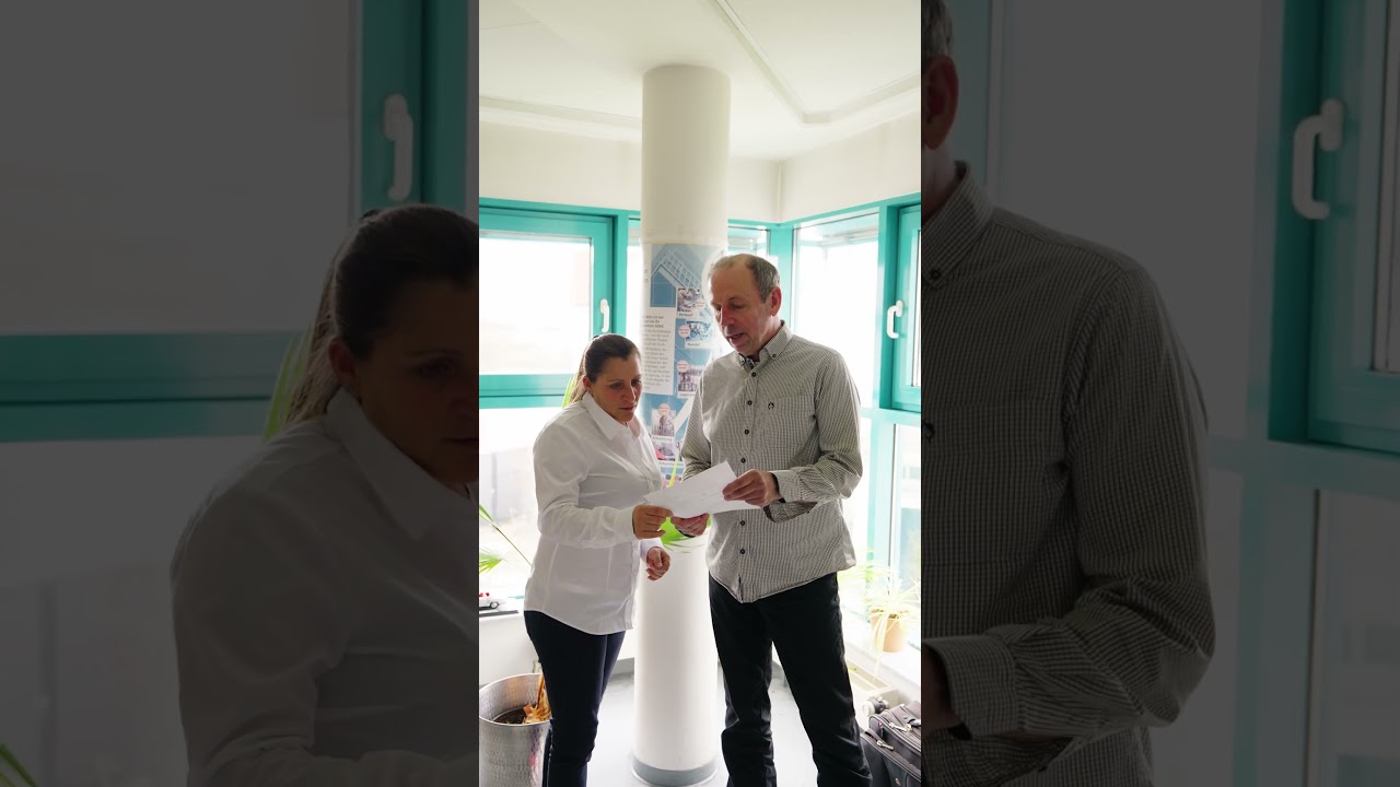 Dieser Alternativtext wurde KI-generiert: Zwei Personen stehen in einem Innenraum vor einer Tür und übergeben sich ein Dokument. Im Hintergrund ist ein Fenster und eine Säule zu sehen.