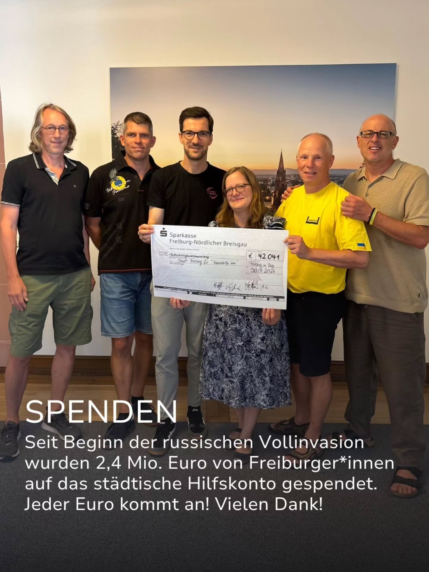 Sechs Personen stehen nebeneinander, zwei davon halten einen großen Scheck der Sparkasse Freiburg-Nördlicher Breisgau über 42.041 Euro. Ein Text-Overlay informiert darüber, dass seit Beginn der russischen Invasion 2,4 Millionen Euro von Freiburger*innen auf ein städtisches Hilfskonto gespendet wurden. (KI-Alternativtext)