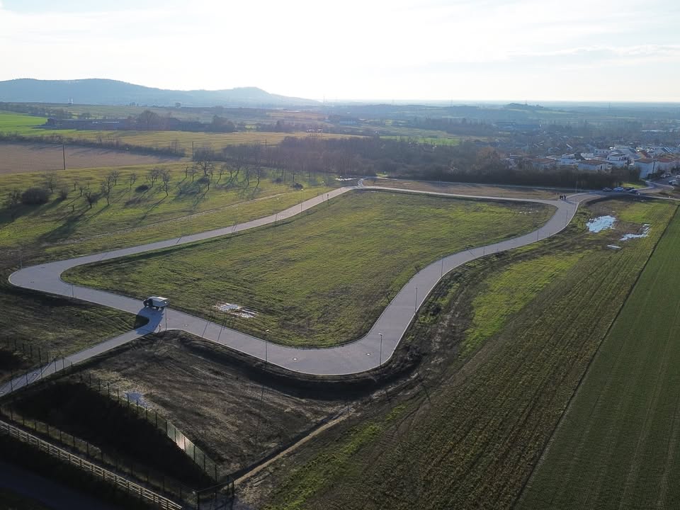 Dieser Alternativtext wurde KI-generiert: Eine Luftaufnahme zeigt eine grasbewachsene Landschaft mit einem Weg, der sich kurvig durch die Fläche schlängelt. Im Hintergrund sind Hügel und einige Gebäude zu sehen.