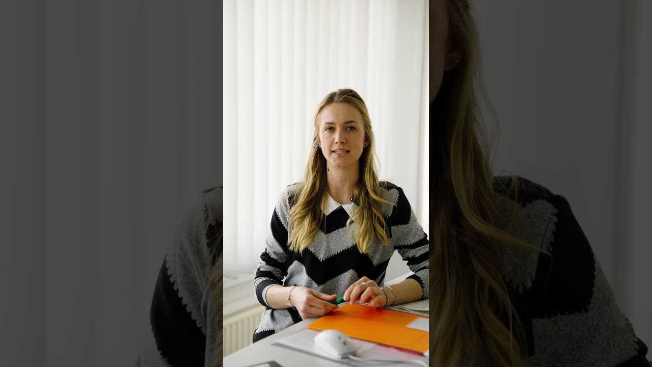 Dieser Alternativtext wurde KI-generiert: Eine Person sitzt an einem Tisch und hält bunte Papierstücke in den Händen. Im Hintergrund ist ein weiterer Teil der Person und ein Fenster zu sehen.