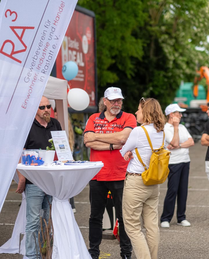 Dieser Alternativtext wurde KI-generiert: Menschen stehen an einem Tisch im Freien, möglicherweise auf einer Veranstaltung. Im Hintergrund ist ein Banner mit Text zu sehen.