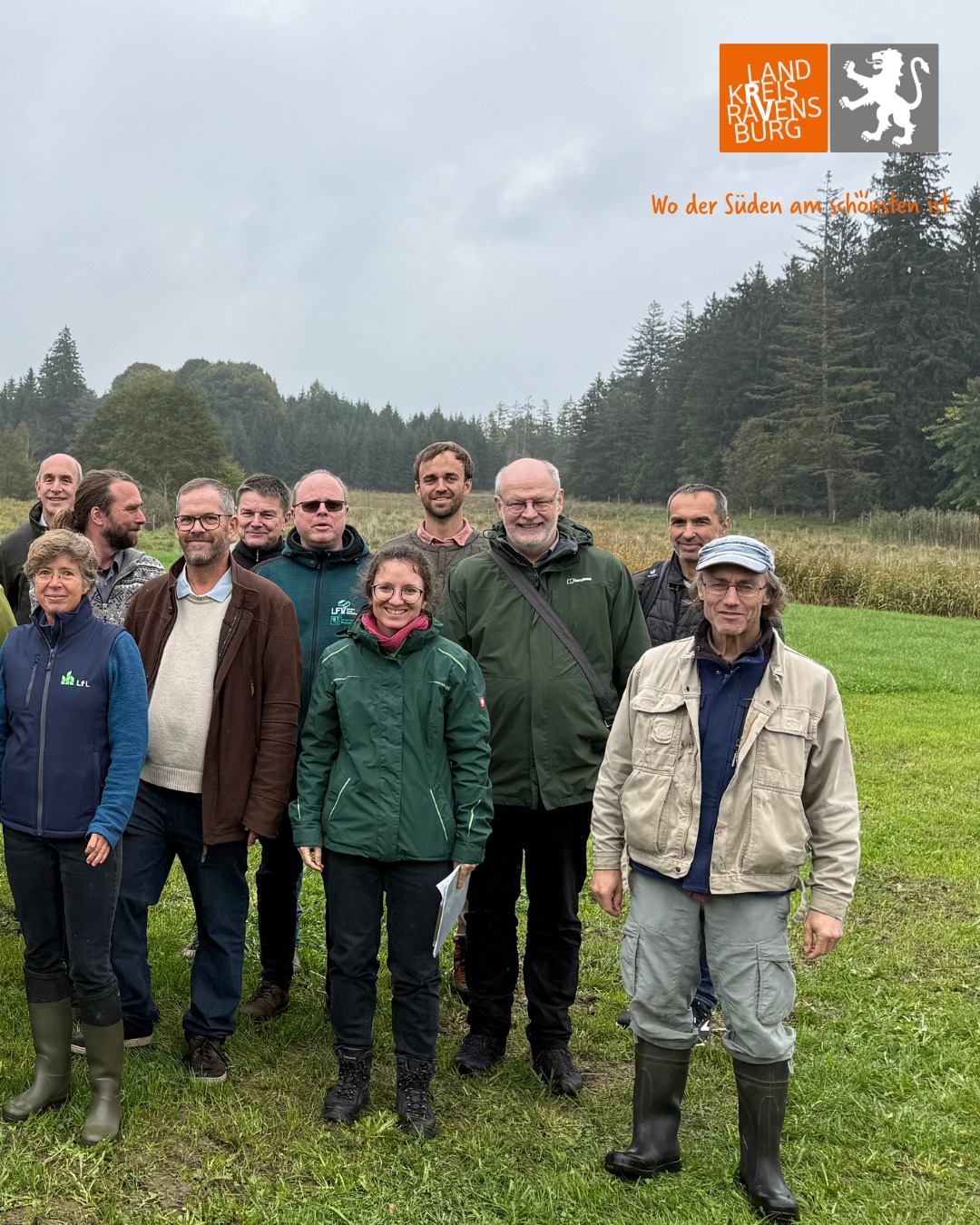 Dieser Alternativtext wurde KI-generiert: Eine Gruppe von Personen steht auf einem Feld vor einem Wald. Im oberen Bildrand befindet sich ein Logo und Text.
