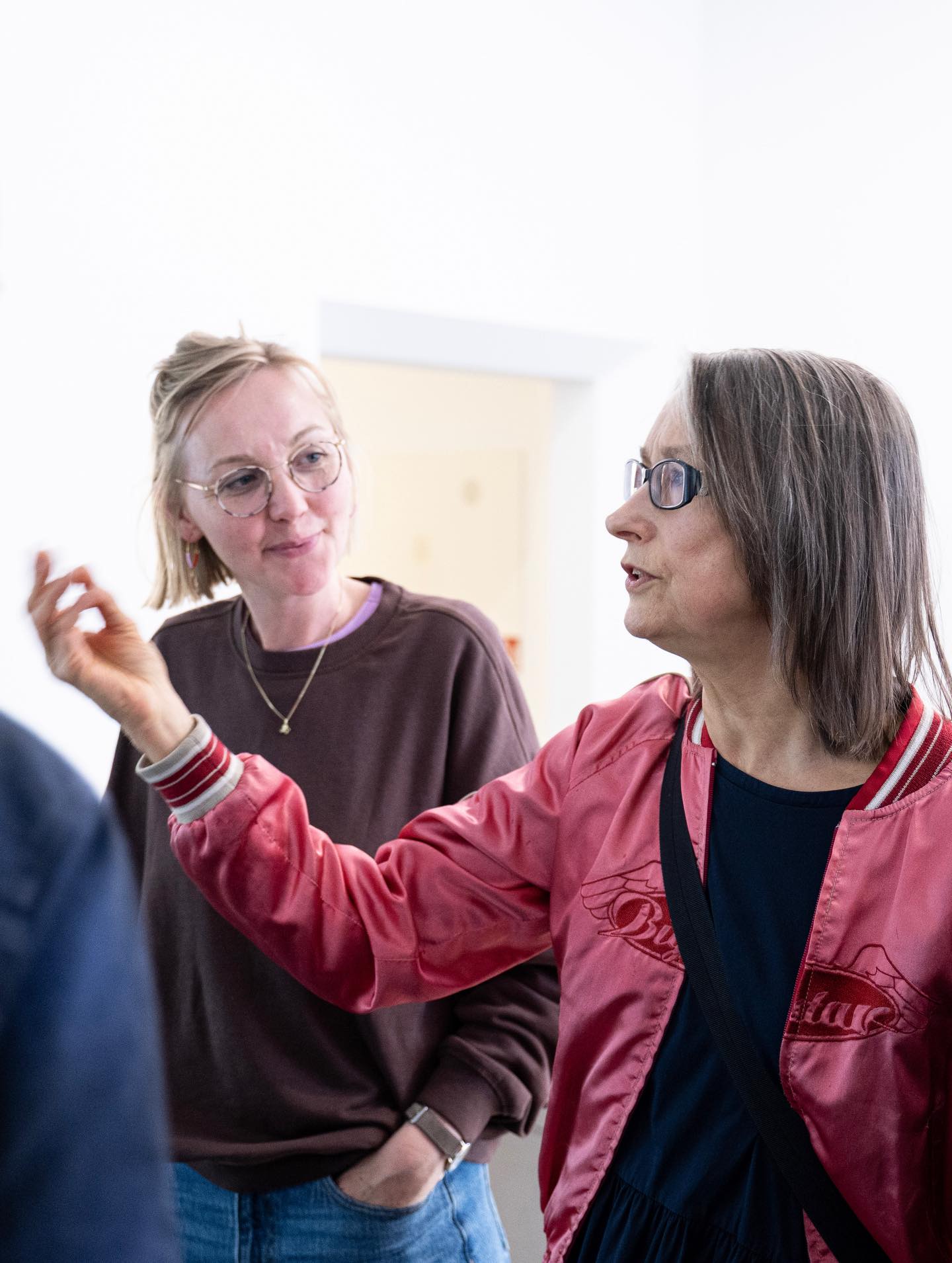 Dieser Alternativtext wurde KI-generiert: Zwei Personen mit Brillen stehen sich zugewandt und scheinen zu sprechen. Im Hintergrund ist ein Teil eines Raumes zu sehen.