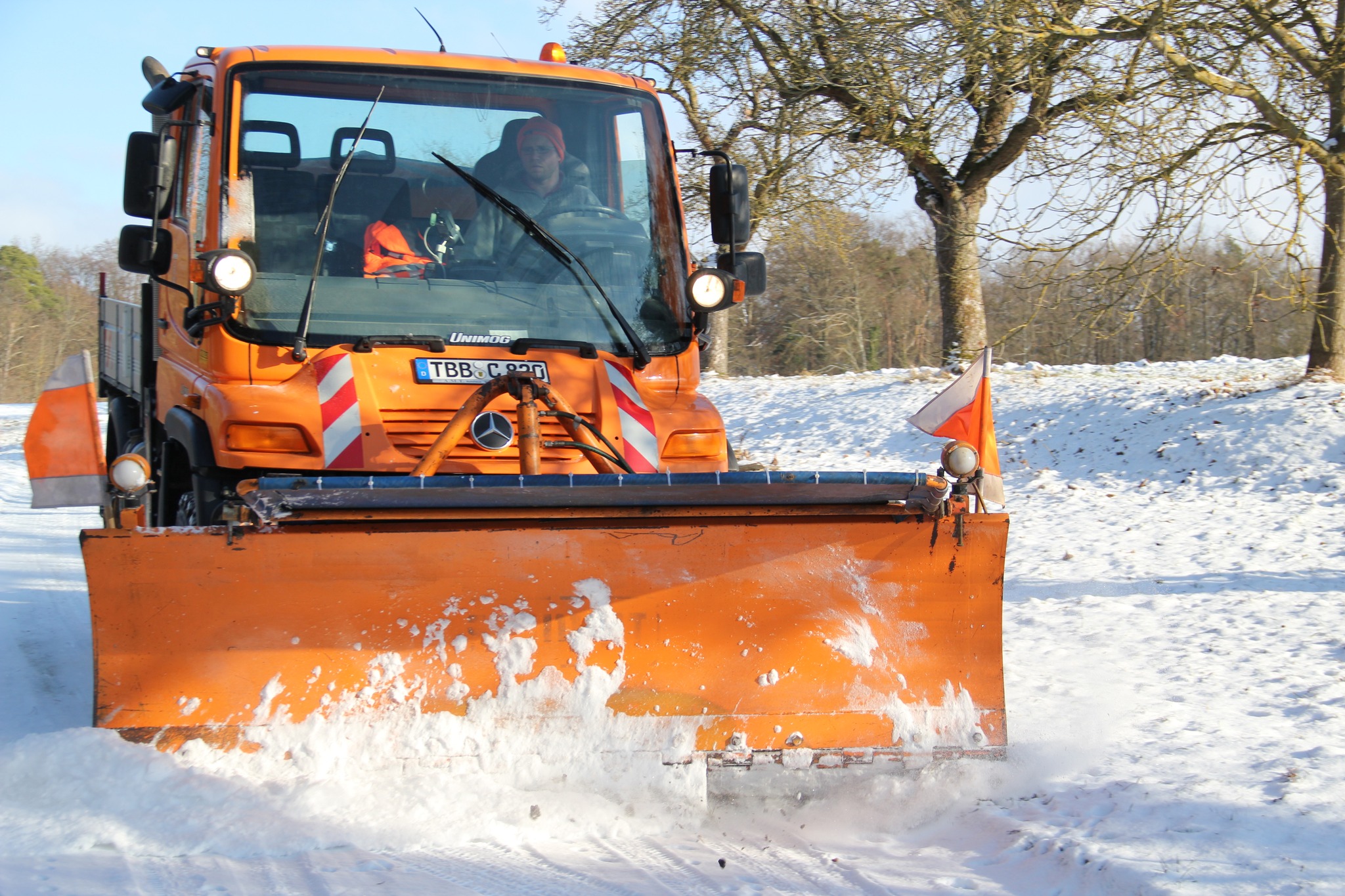 Dieser Alternativtext wurde KI-generiert: Ein orangefarbenes Fahrzeug mit Schneeschild räumt Schnee von einem Feldweg. Im Fahrgastraum ist eine Person zu erkennen.