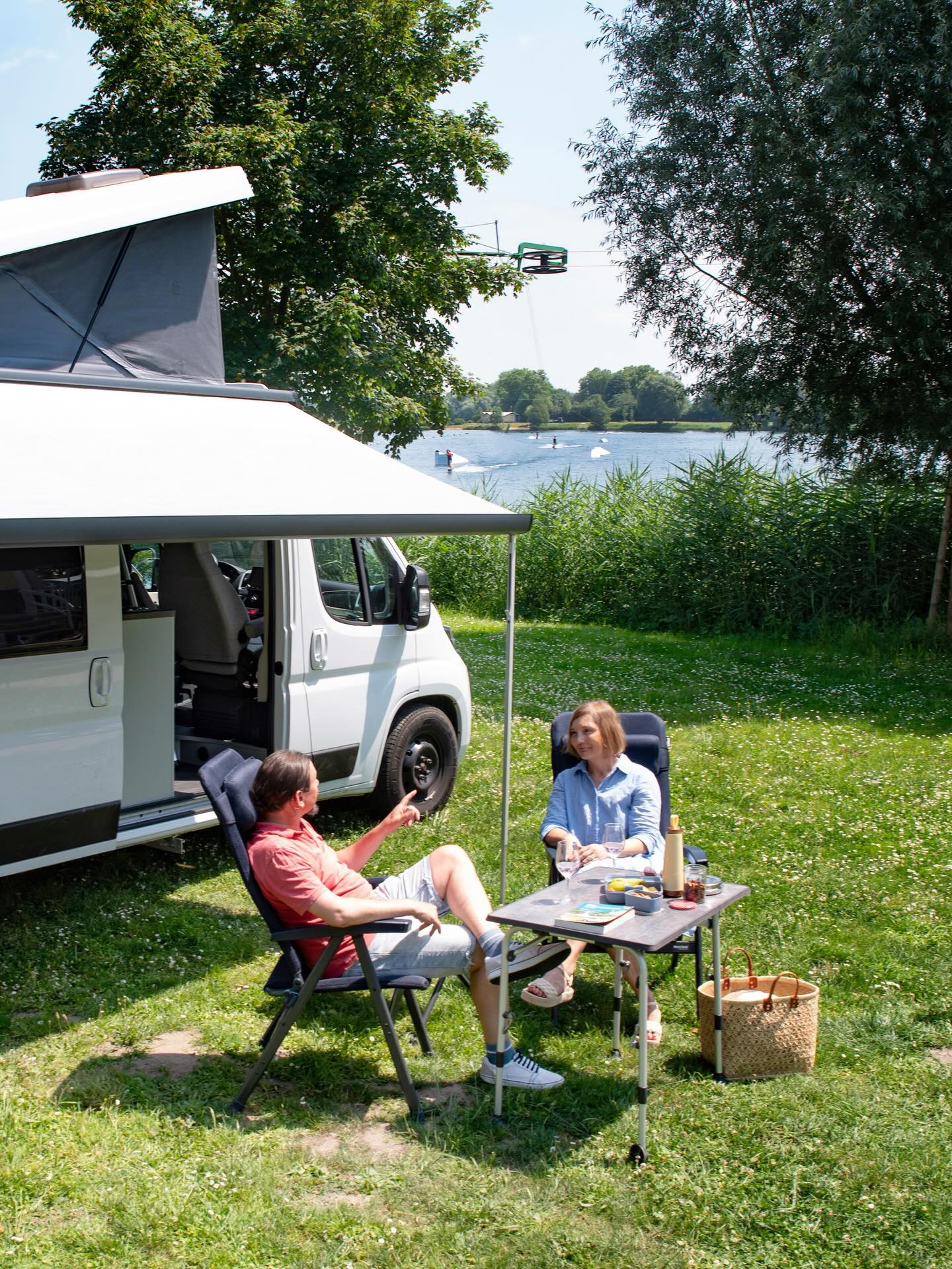 Zwei Personen sitzen auf Campingstühlen unter der Markise eines weißen Wohnmobils mit Dachzelt und unterhalten sich. Auf dem Tisch zwischen ihnen stehen Gläser und Lebensmittel. Im Hintergrund ist ein See mit Wassersportlern zu sehen. (KI-Alternativtext)
