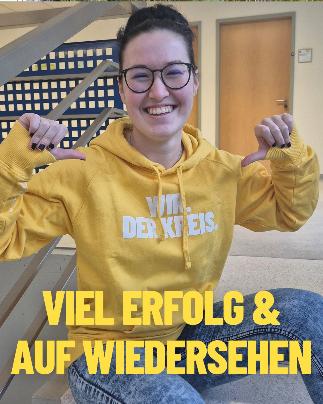 Dieser Alternativtext wurde KI-generiert: Eine Person in einem gelben Pullover lächelt in die Kamera und zeigt mit beiden Händen nach oben. Im Hintergrund ist ein Treppengeländer und ein Schild mit einem Spruch zu sehen.