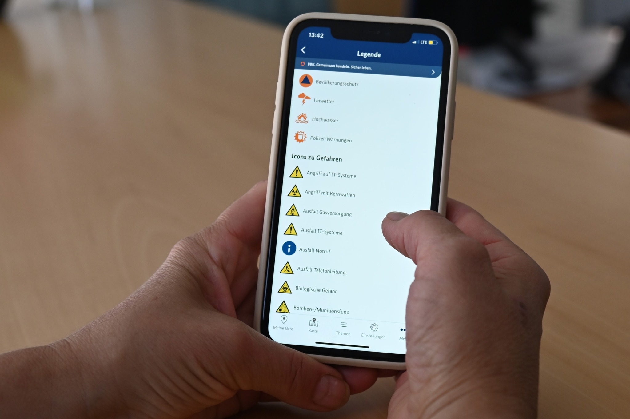 Zwei Hände halten ein Smartphone, das eine App-Legende mit verschiedenen Warnsymbolen anzeigt. Die Liste umfasst Kategorien wie Bevölkerungsschutz, Unwetter und Hochwasser sowie eine Sektion "Icons zu Gefahren" mit Einträgen wie Angriff auf IT-Systeme, Kernwaffen, Gasausfall und biologische Gefahr. Am unteren Rand ist eine Navigationsleiste mit den Punkten Meine Orte, Karte, Themen und Einstellungen sichtbar. (KI-Alternativtext)