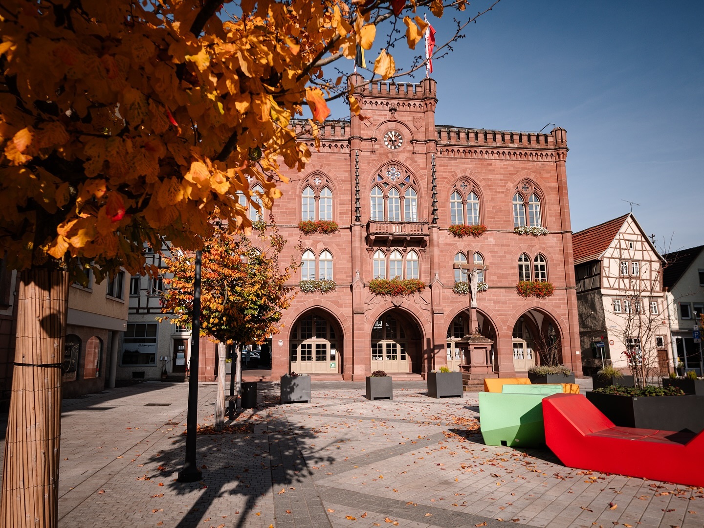 Ein Rathaus aus rotem Sandstein steht auf einem Platz mit Bäumen in Herbstfarben. Moderne Sitzgelegenheiten stehen vor dem Gebäude.